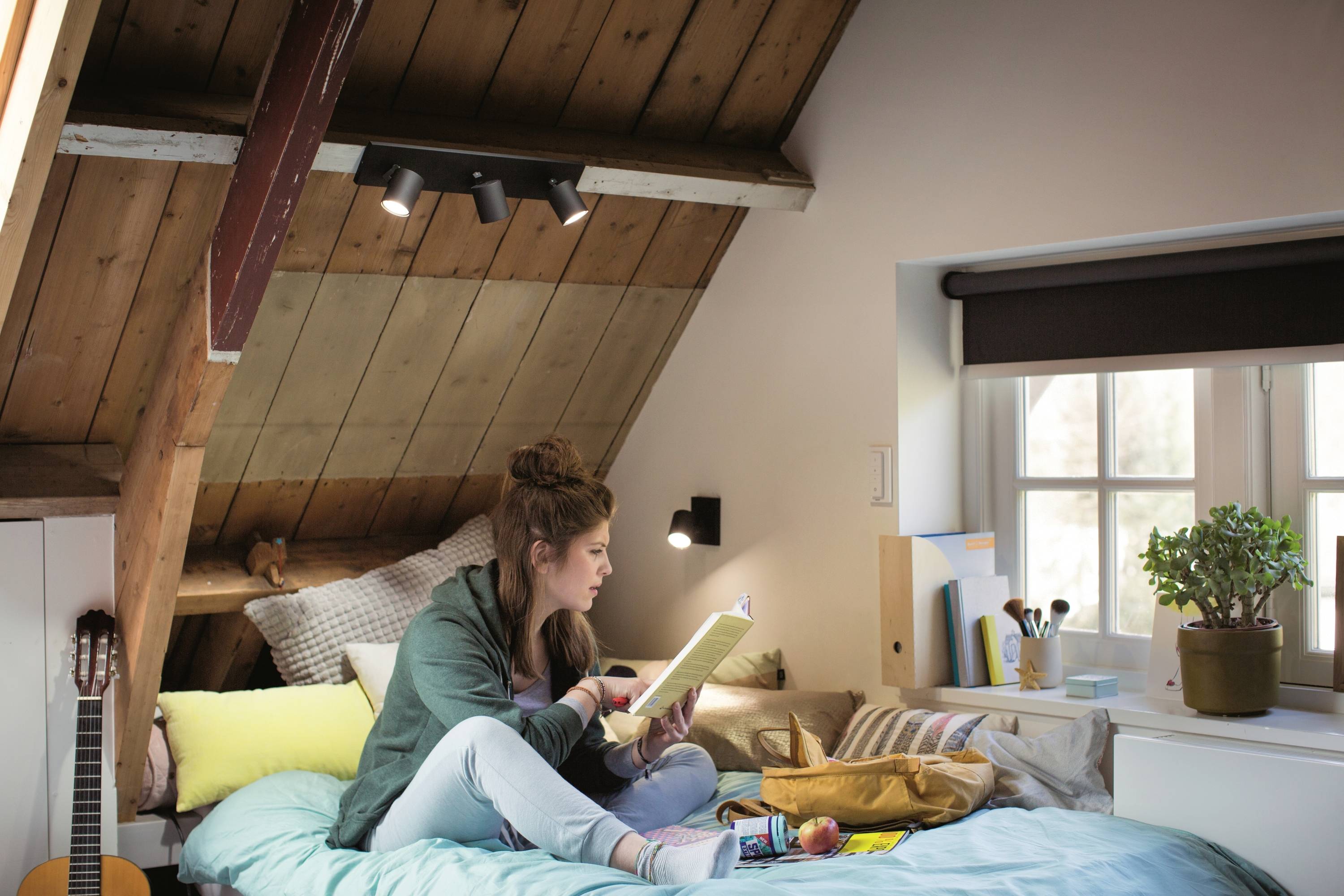 A young woman sits on a bed beneath a skylight, reading a notebook. Books, a laptop, and a guitar are lying beside her.