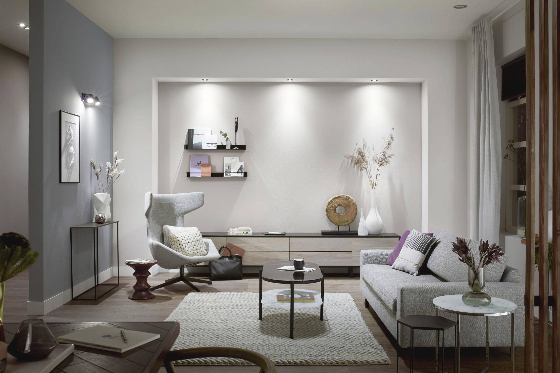 Modern living room with light walls, grey sofa, armchair and shelves on the wall. Decoration including vases, books and a lamp.