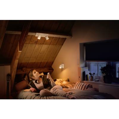 A woman is lying on a bed in a cosily lit attic room, holding a remote control while looking upwards.