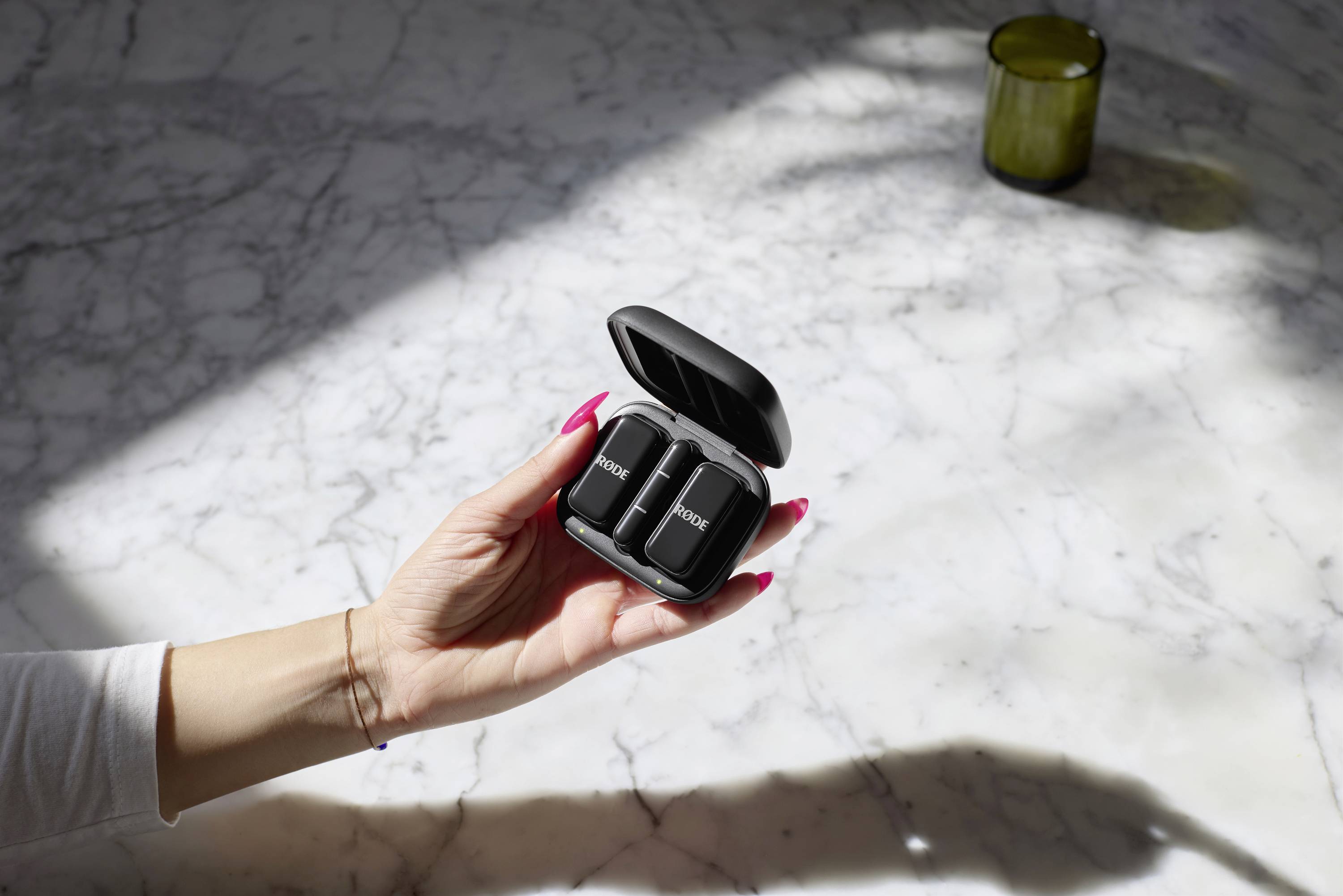 A person is holding an open earphone case with two earbuds inside, placed on a marble table with sunlight in the background.