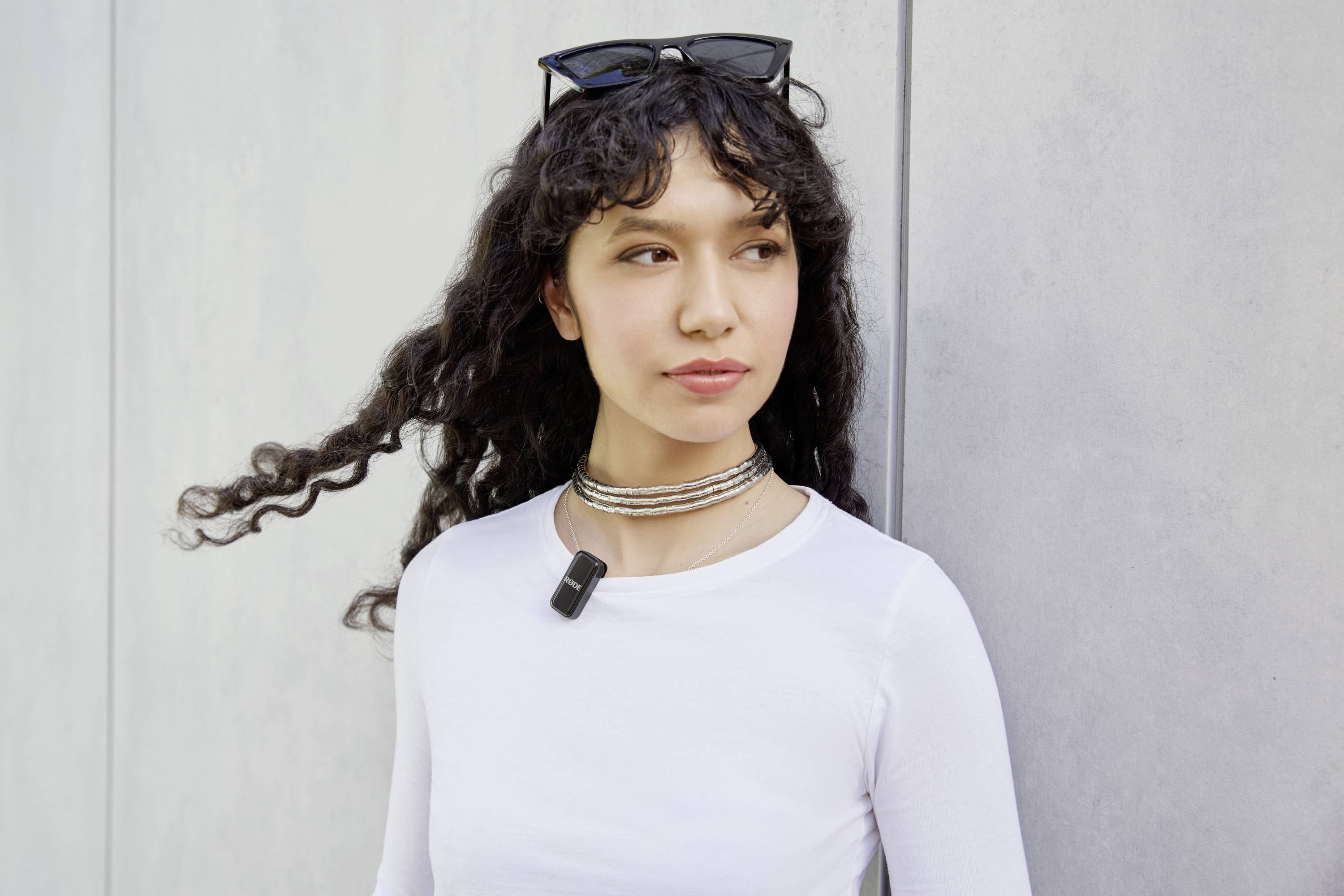 A woman with curly hair and a white top is leaning against a wall. She is wearing glasses on her head and has a pendant around her neck.