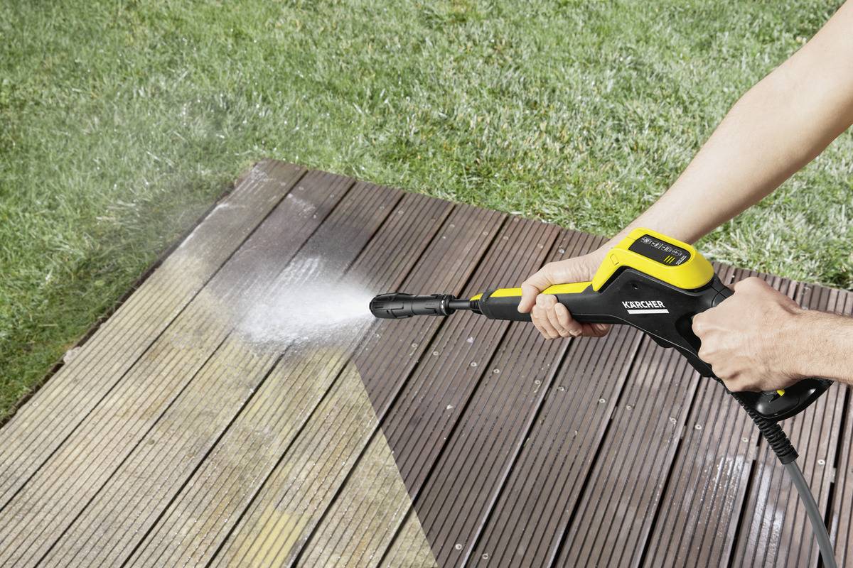 A person is cleaning a wooden terrace with a pressure washer. On the left is a clean sprayed area, on the right is still uncleaned.