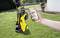 'A yellow pressure washer is standing on a lawn. A hand is holding a smartphone displaying an advert of the cleaner.'