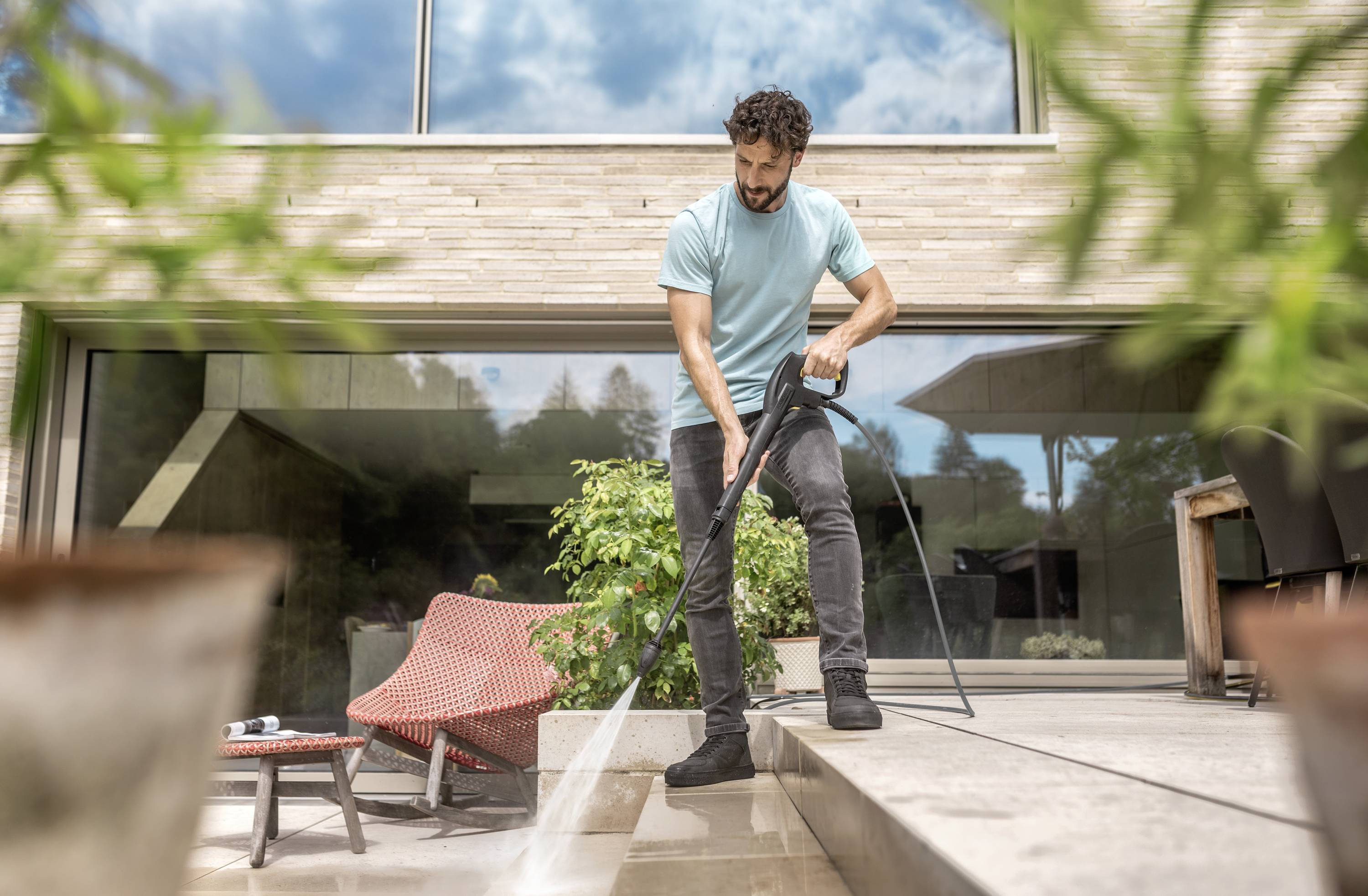 A man is cleaning the terrace of a modern house with a pressure washer. Large windows and plants can be seen in the background.