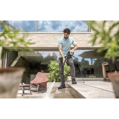A man is cleaning the terrace of a modern house with a pressure washer. Large windows and plants can be seen in the background.