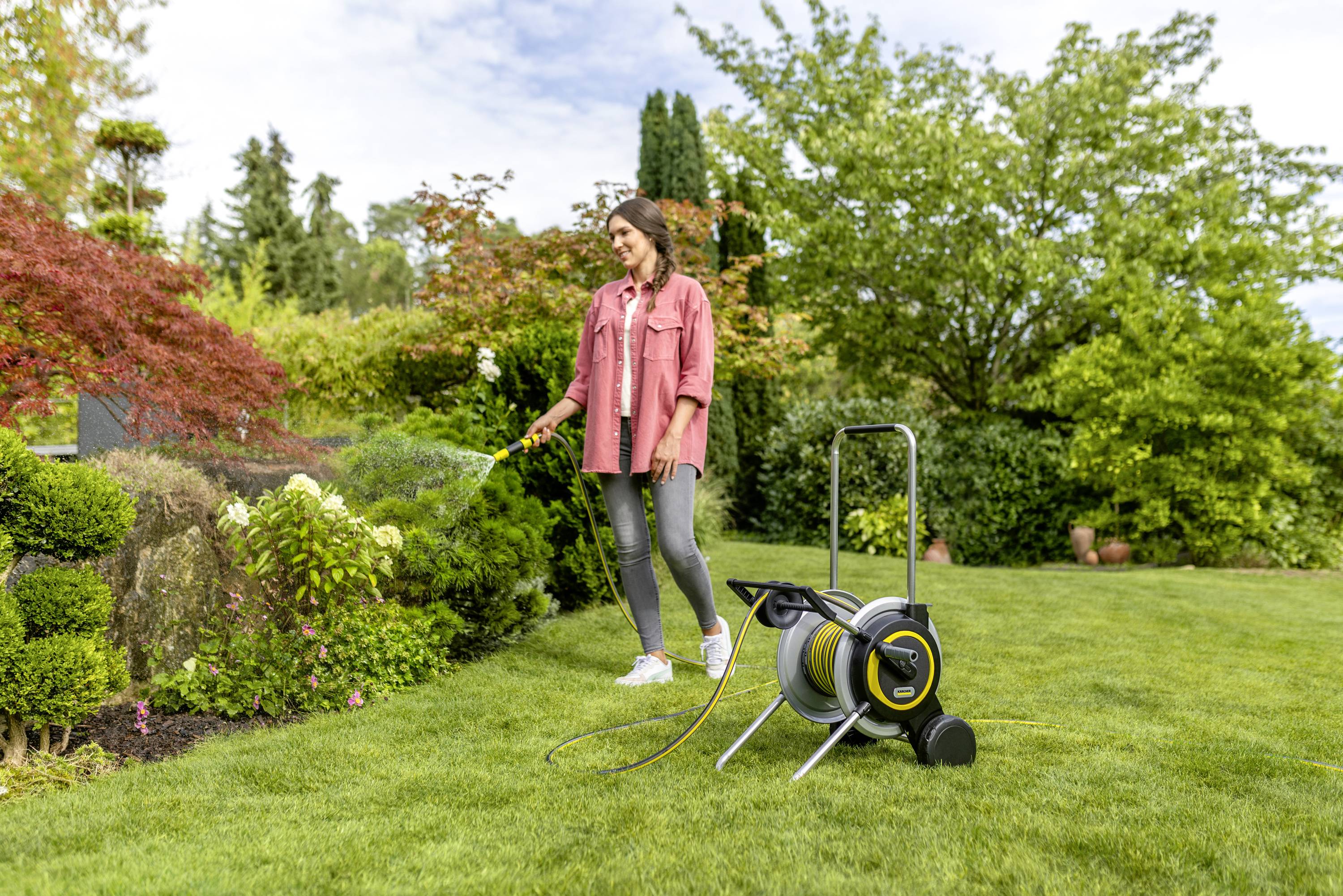 A woman is watering plants with a hosepipe in a well-maintained garden. A hose reel stands on the lawn.
