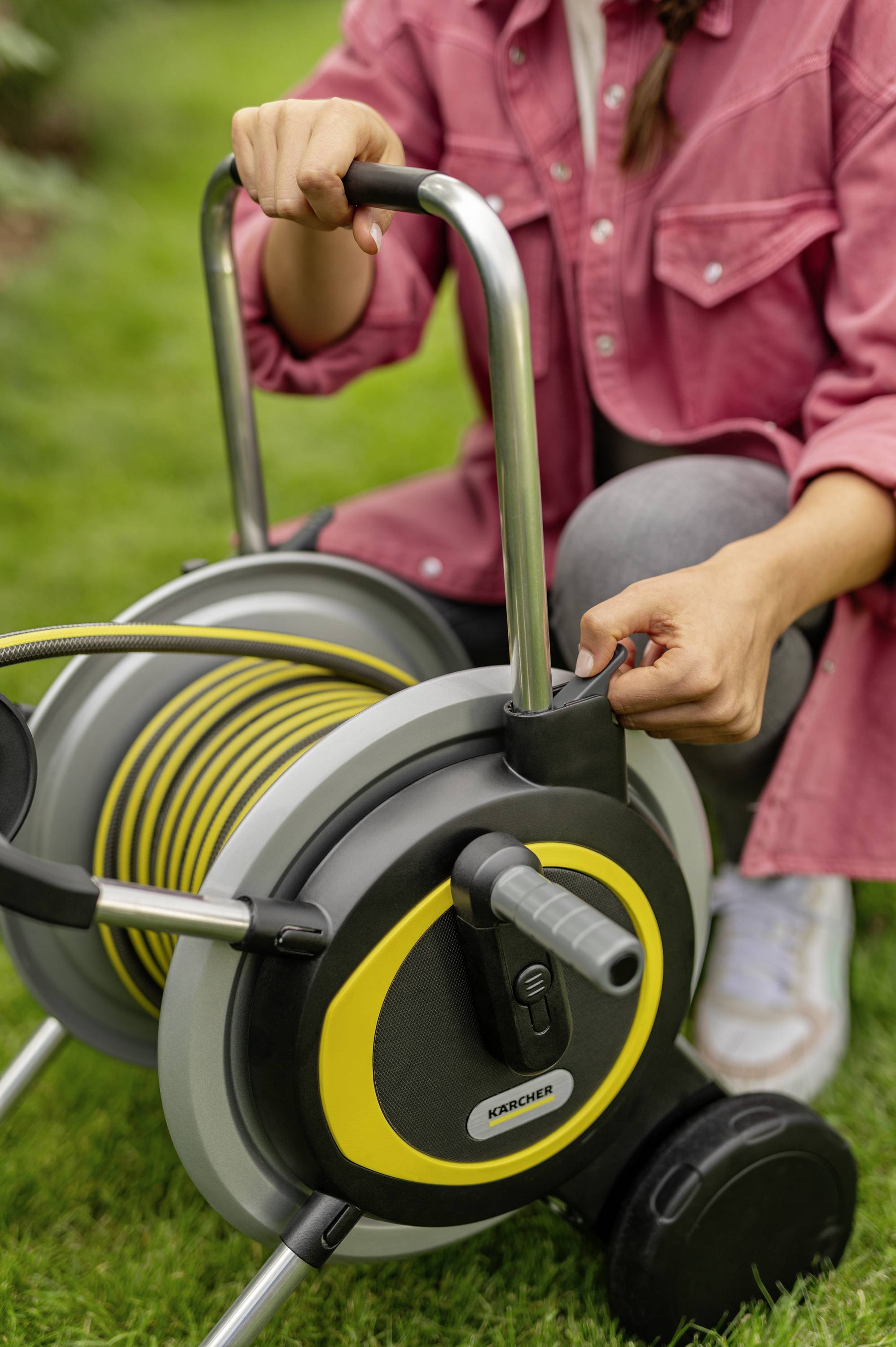 A person in the garden is holding a hose reel with a yellow hose and black wheels, ready to water the lawn.