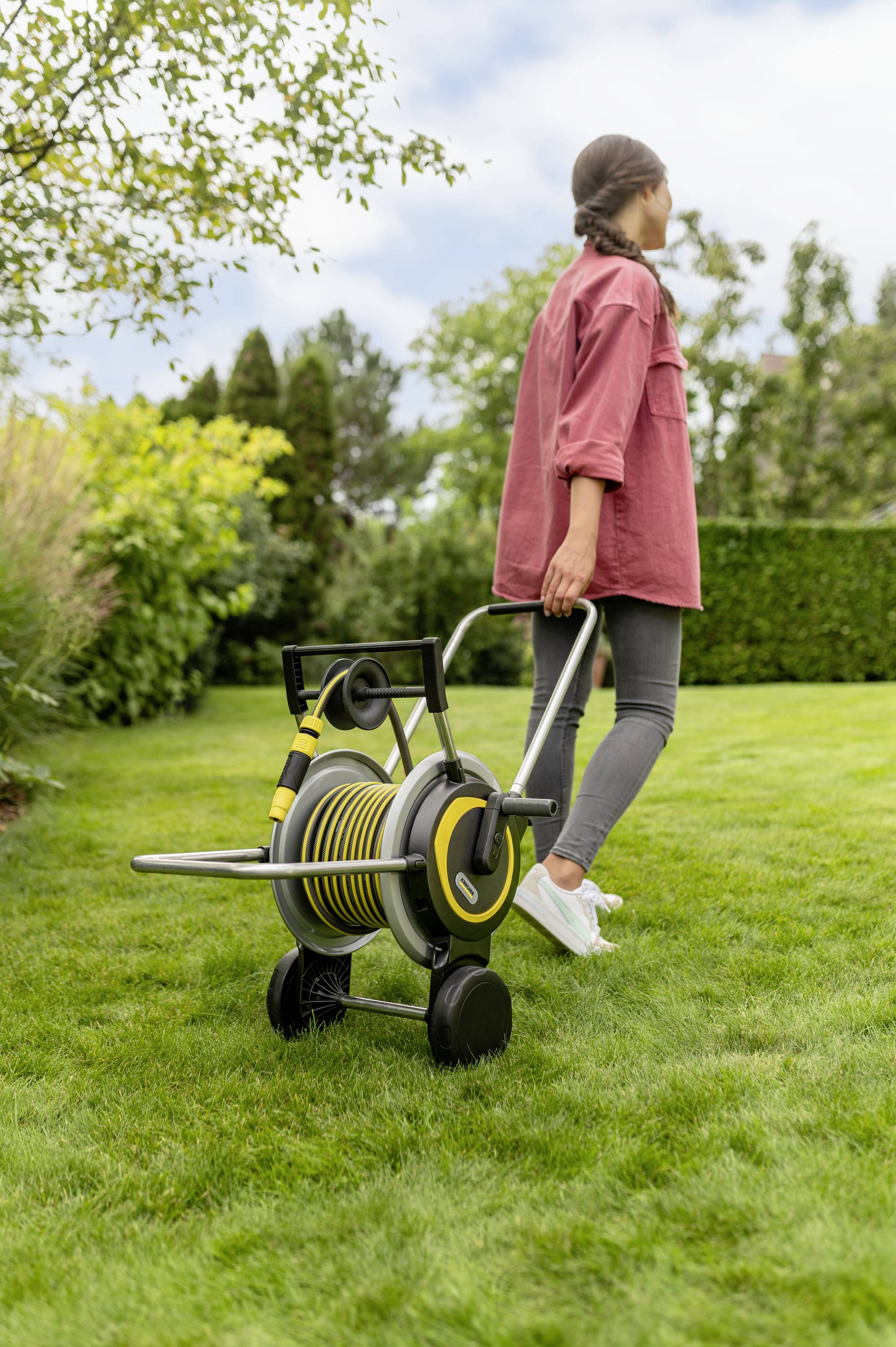 A person is pulling a garden hose reel across a lawn. The garden in the background is green and well-maintained.