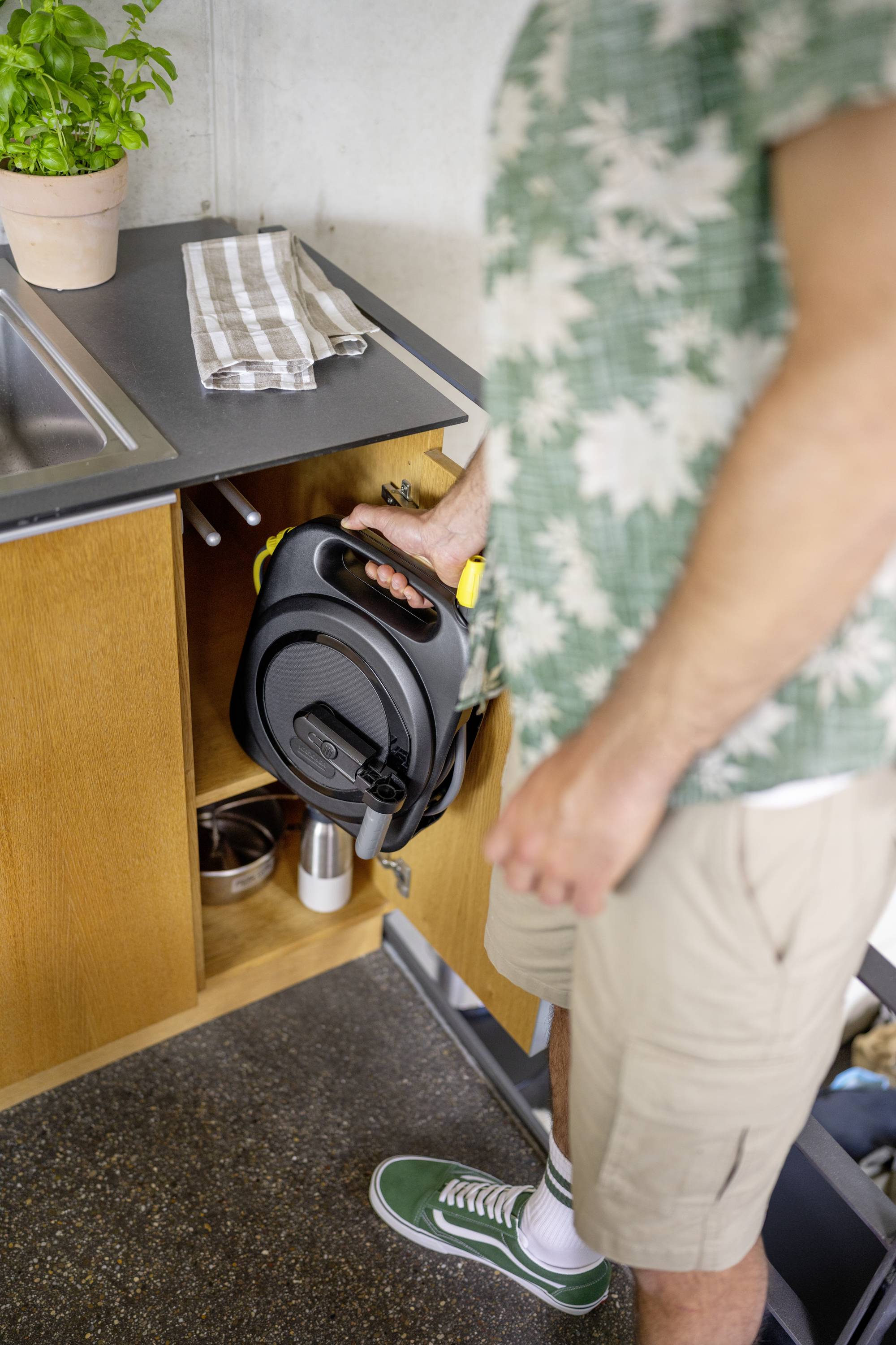 A person wearing summer clothing opens a lower kitchen cupboard. A rolled-up extension cable reel is lying inside the cupboard.
