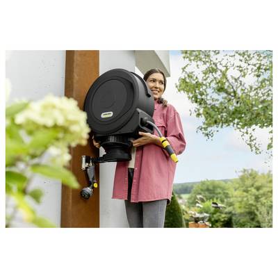 A person in a pink shirt installs a black garden hose reel on an outdoor wall. A blooming plant and trees are visible in the background.