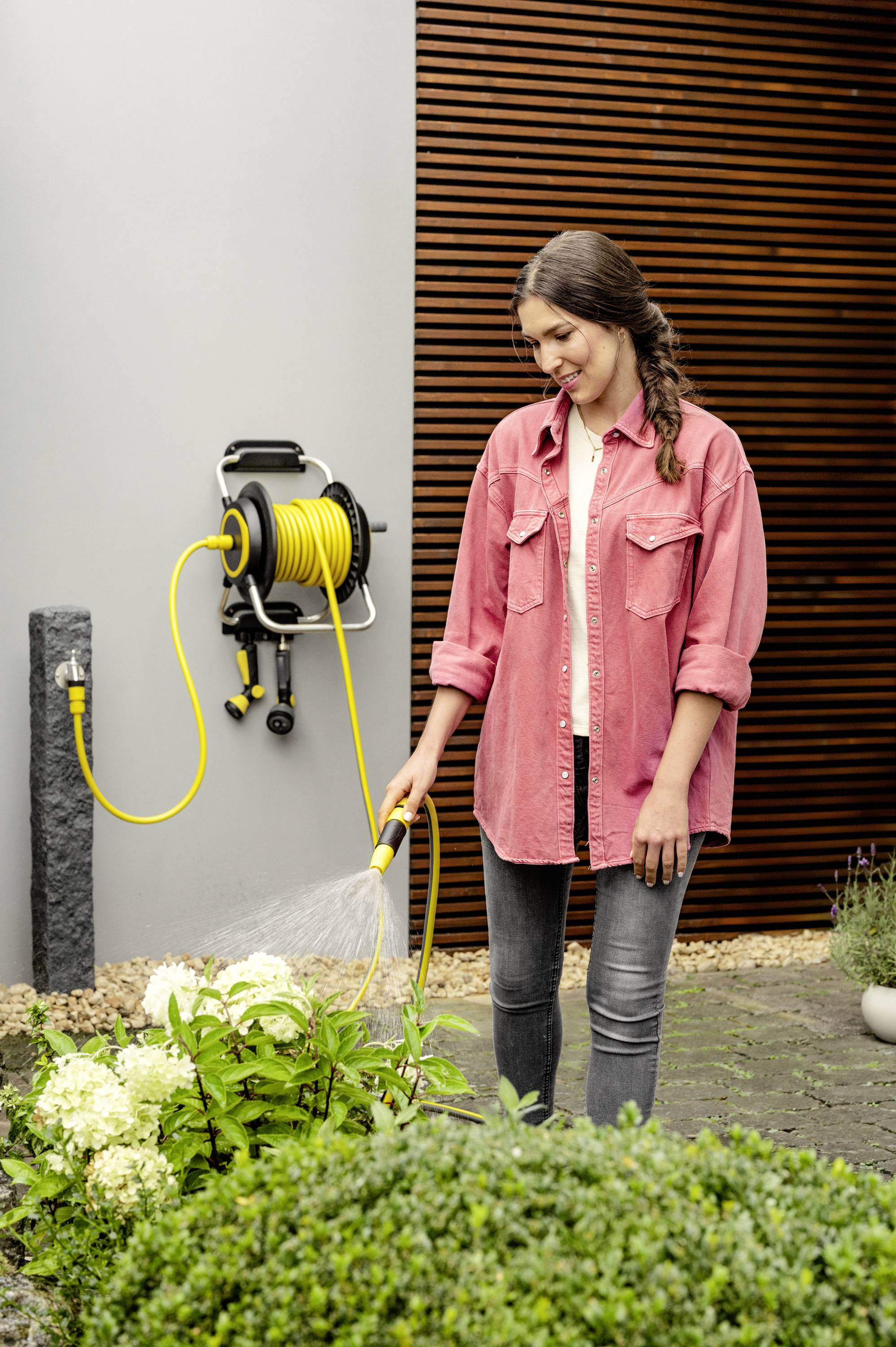 A woman in a pink jacket is watering plants with a yellow garden hose. In the background, a wall with wooden panelling.