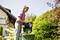 A woman empties the grass catcher of a lawnmower into a rubbish bin in the garden, surrounded by green plants and a house in the background.