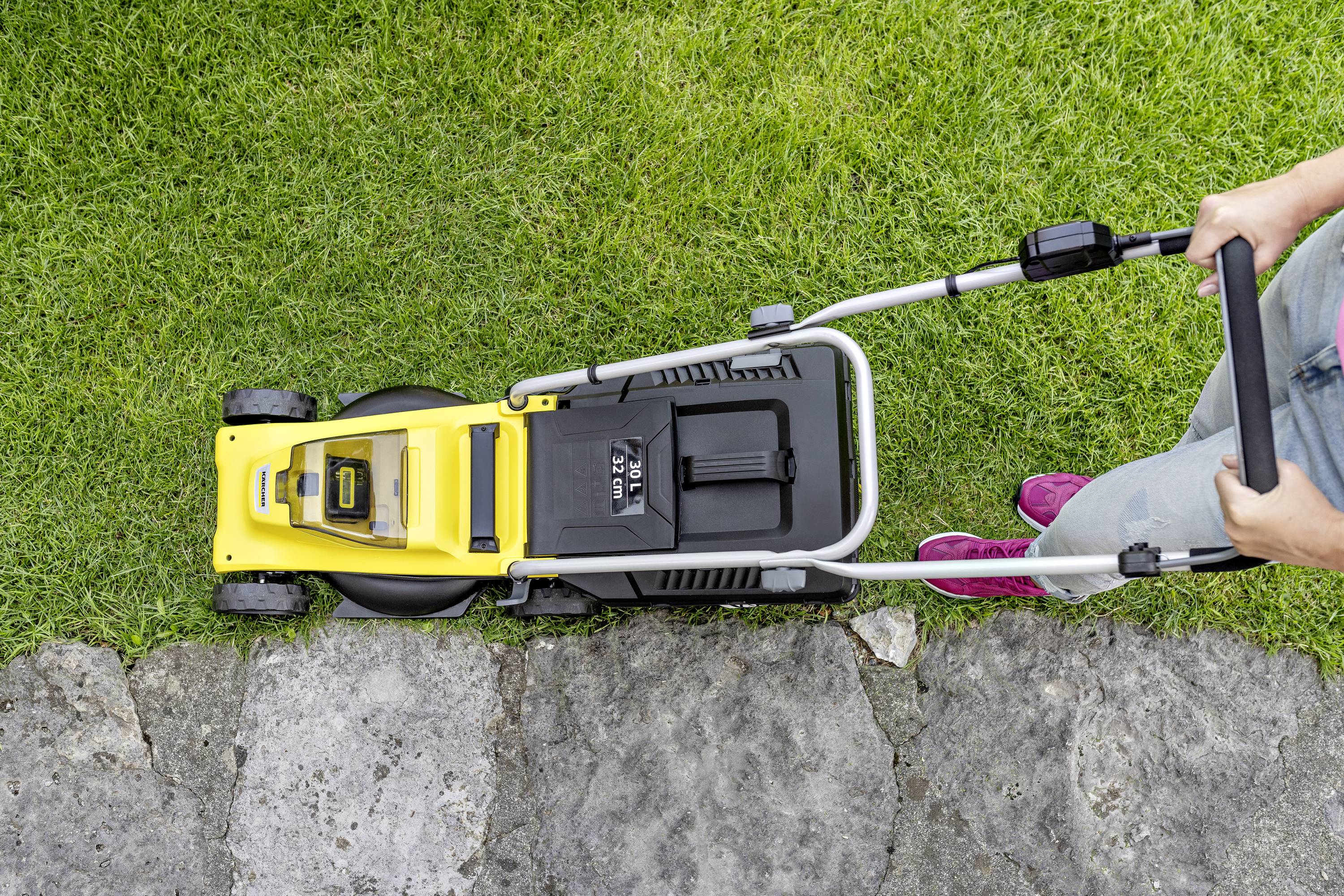A person is pushing a yellow lawnmower along a green lawn and stops at the edge of a paved path.