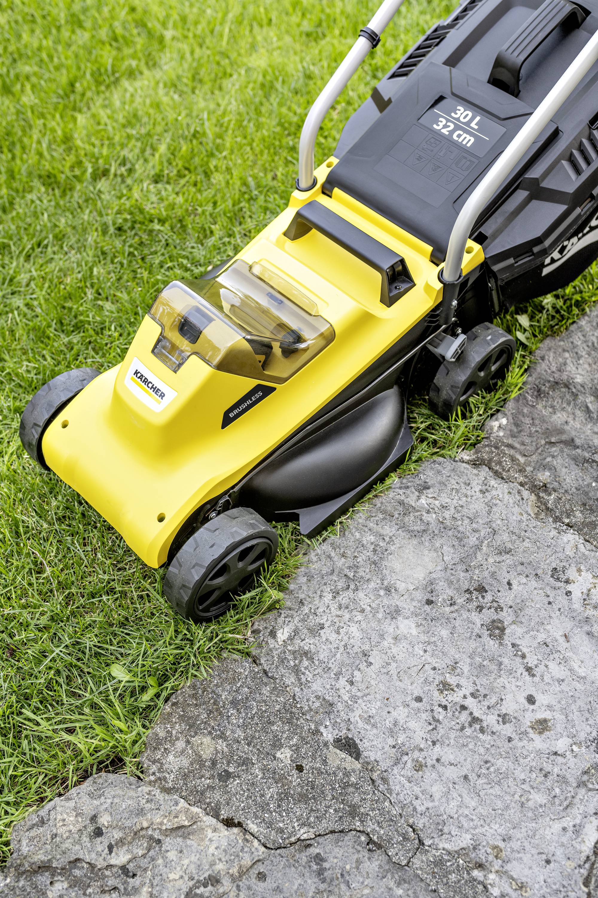 A yellow lawnmower is cutting grass along a paved path. The mower has a transparent collection bag and black wheels.