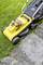 A yellow lawnmower is cutting grass along a paved path. The mower has a transparent collection bag and black wheels.