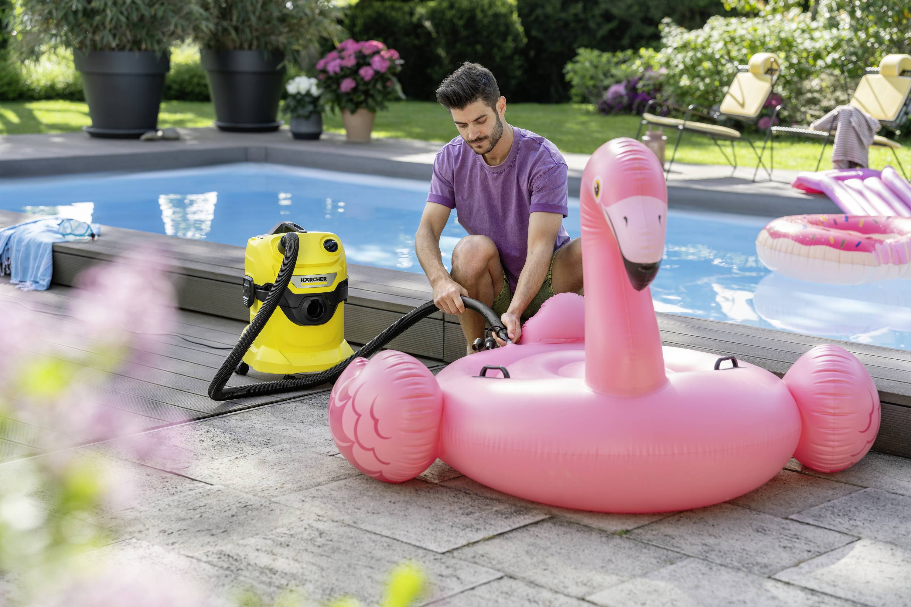 A man is inflating a large, pink flamingo pool float with a yellow device by the pool. Plants are visible in the background.