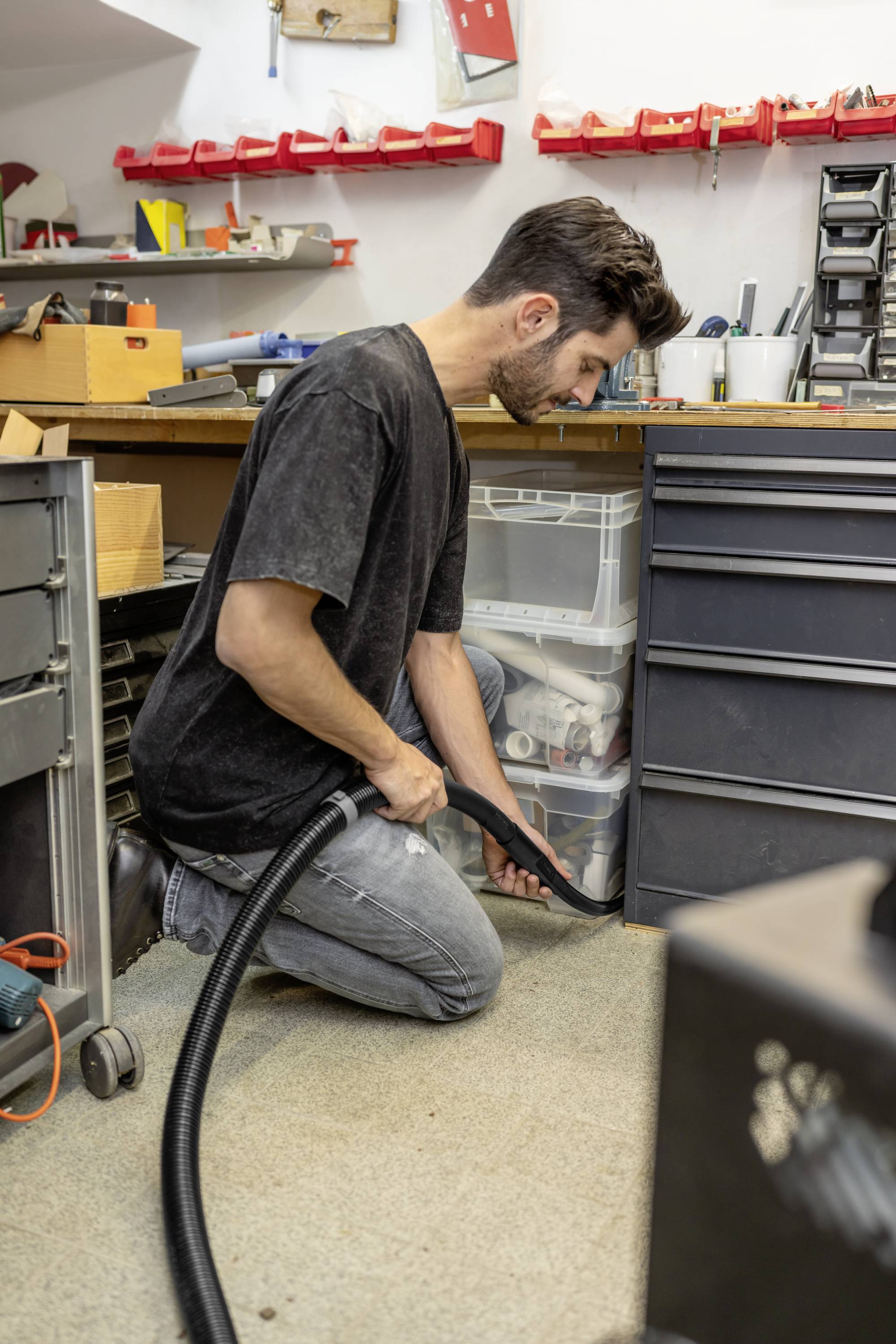 Young man kneels on a workshop floor and vacuums. Tools and shelves are visible in the background.