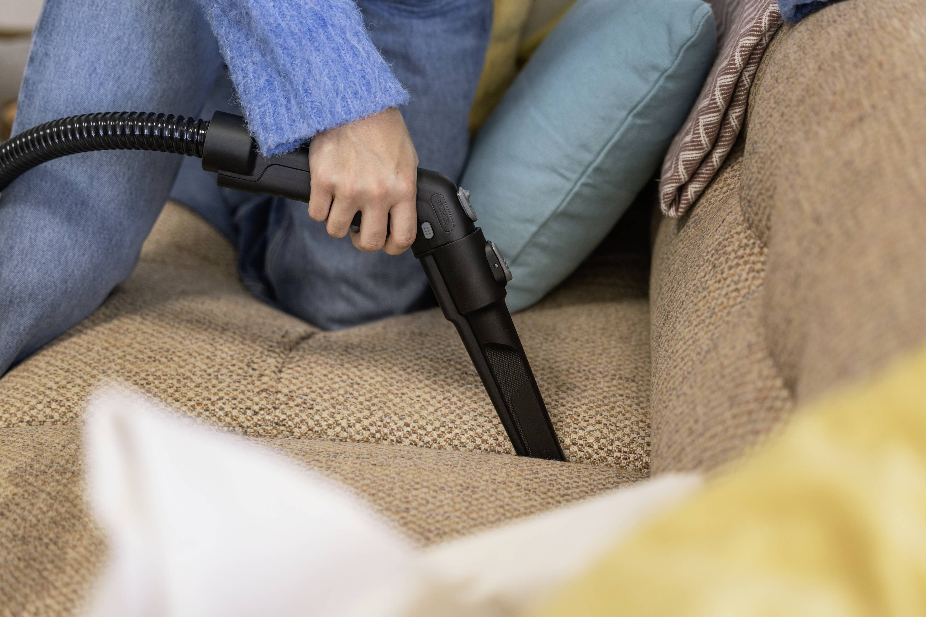 A person is vacuuming a fabric sofa with a vacuum cleaner attachment. Colourful cushions can be seen in the background.