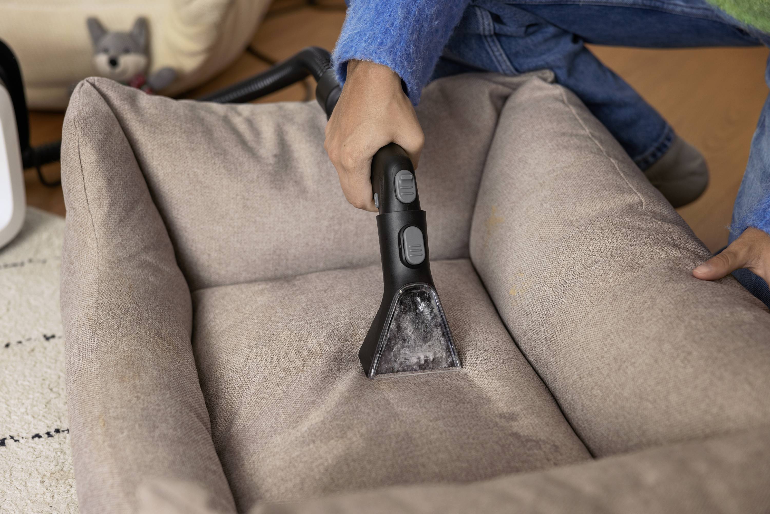 A person is cleaning a brown dog bed with a handheld vacuum cleaner. A dog toy can be seen in the background.