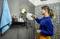 A woman is cleaning a shower screen in a bathroom using a steam cleaner. Various toiletry products are standing on the shelf.