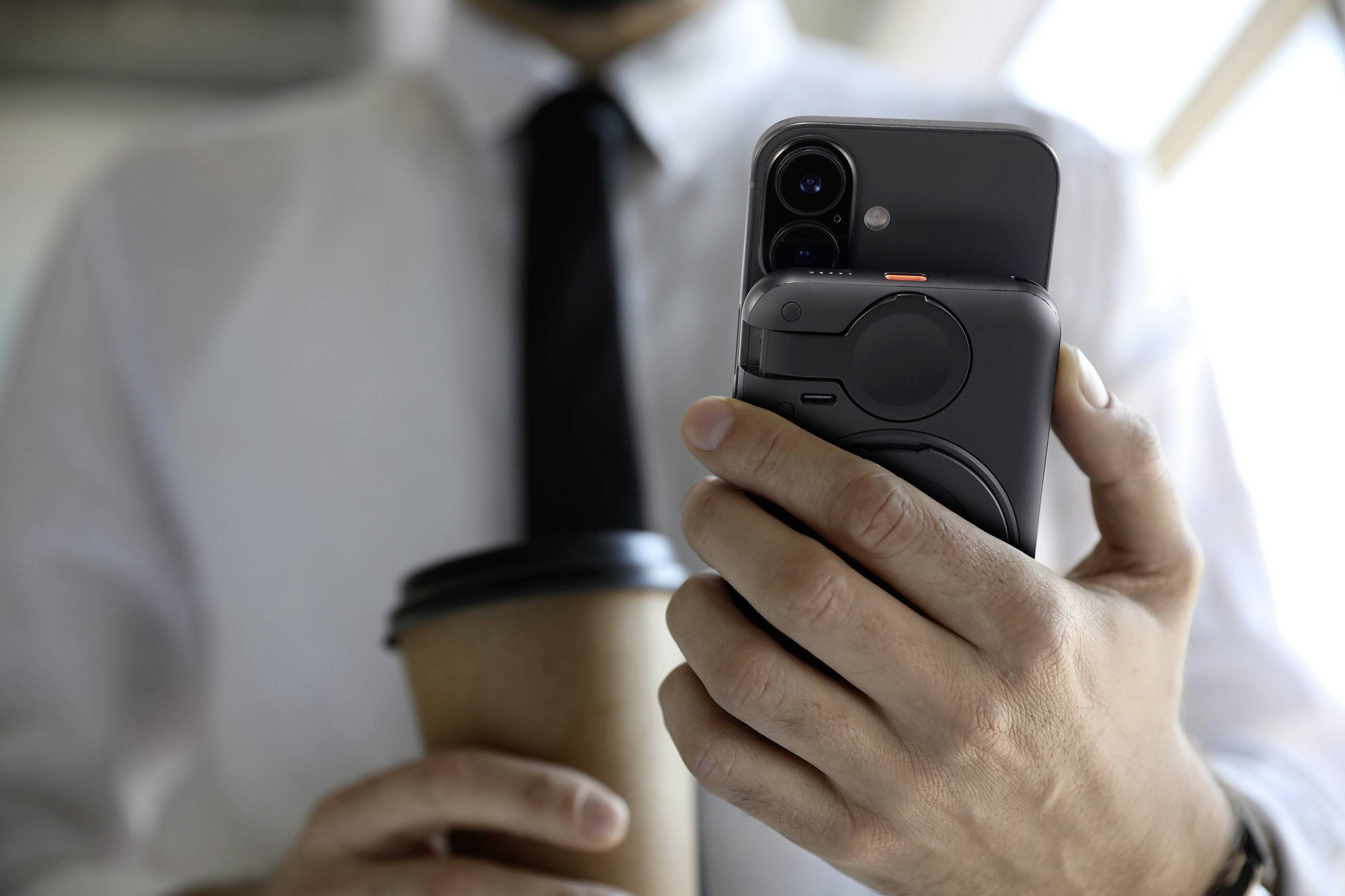 A person is holding a smartphone in their right hand and a paper cup in their left hand, wearing a white shirt and a black tie.