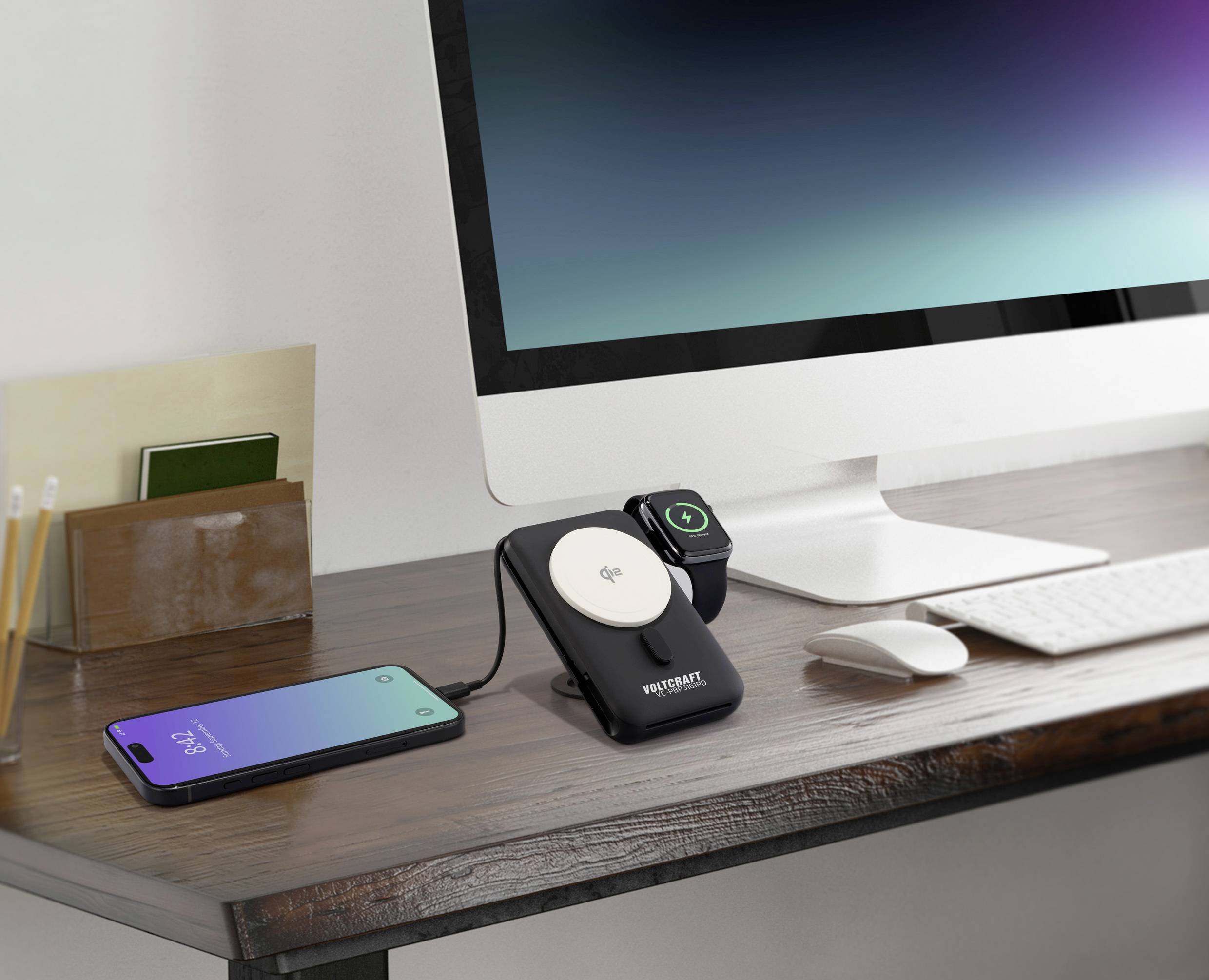 A desk with a computer, a wireless charger with a smartphone and a smartwatch resting on it. A monitor is in the background.