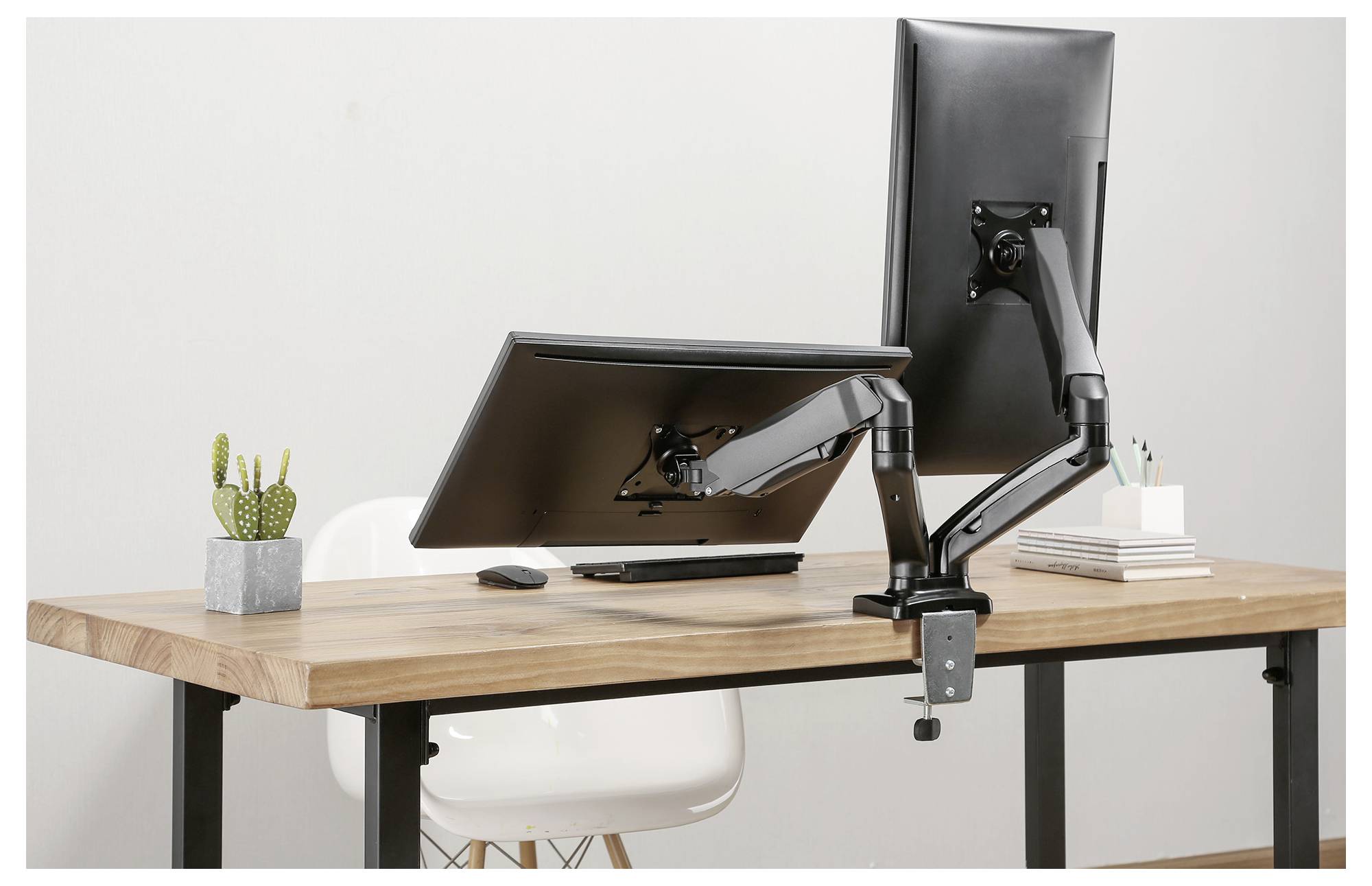 Two monitors are mounted on adjustable arms on a desk. Beside them are a cactus, books, and a mug.