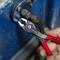 A hand firmly grips a pair of pliers working on a blue metal valve. The pliers have red handles and are grasping a screw.