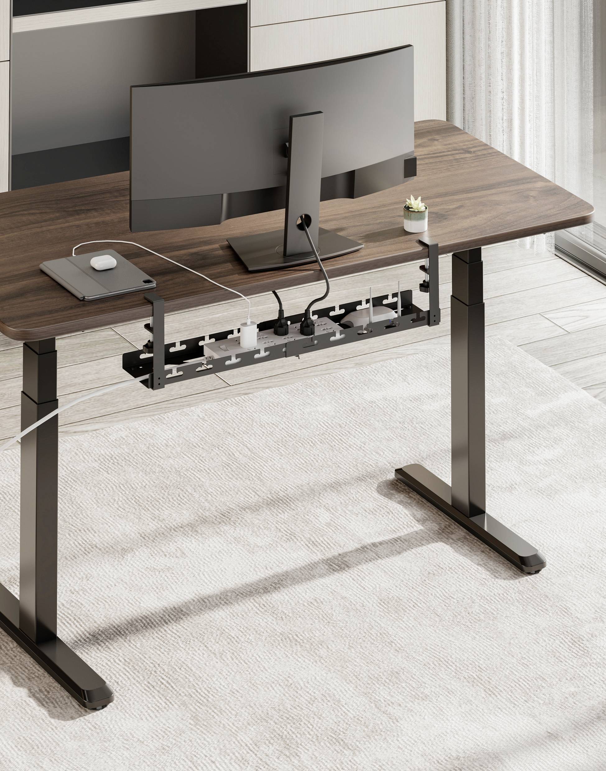 Height-adjustable desk with a wooden surface, featuring a monitor and laptop. Cable management system underneath the desk organises cables.
