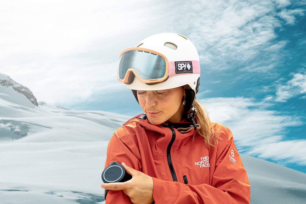 A person in a red jacket and white helmet stands in a snow-covered mountain landscape, looking down at a device on their arm.