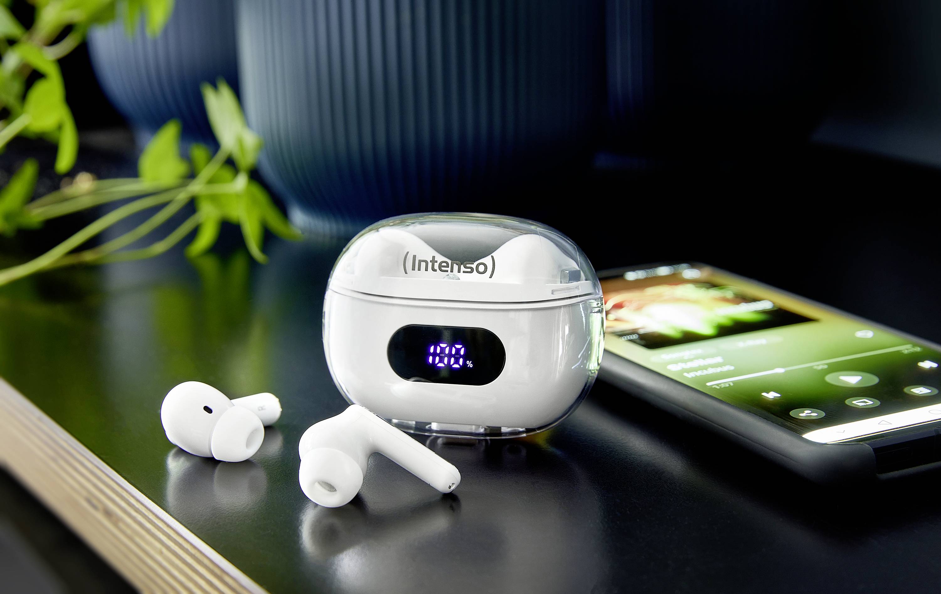 Wireless earbuds with charging case on a table next to a smartphone playing music. A plant is situated in the background.