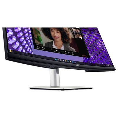 A curved monitor displays a video conference with multiple participants. A woman is speaking, with another display showing light effects in the background.