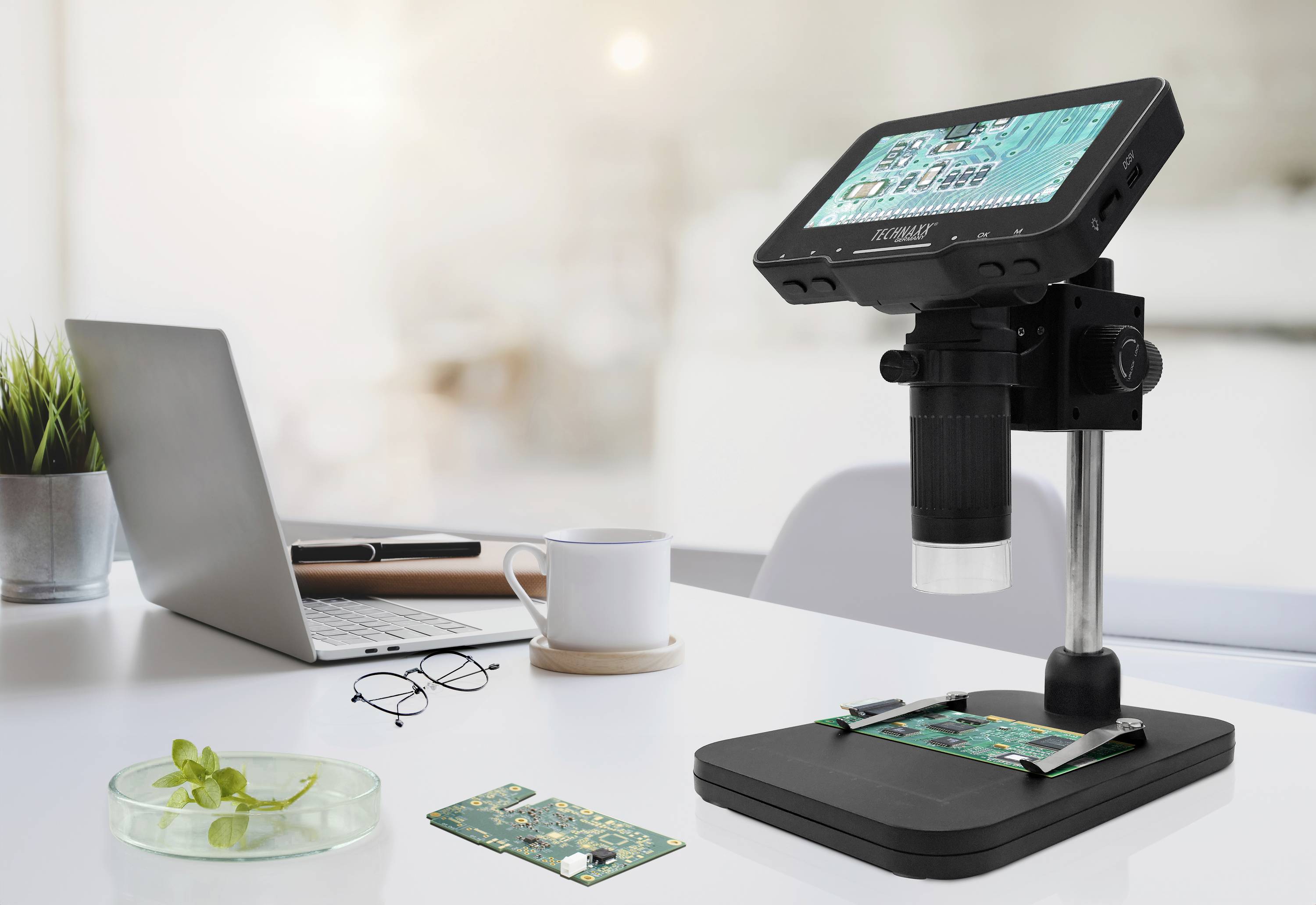 Microscope with display showing circuit boards, sits on a desk with a laptop, glasses, coffee mug and plant in the office.