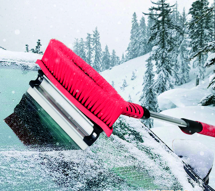 A hand is holding a red ice scraper with a brush, removing snow from a car windscreen. Snow-covered trees and mountains are visible in the background.