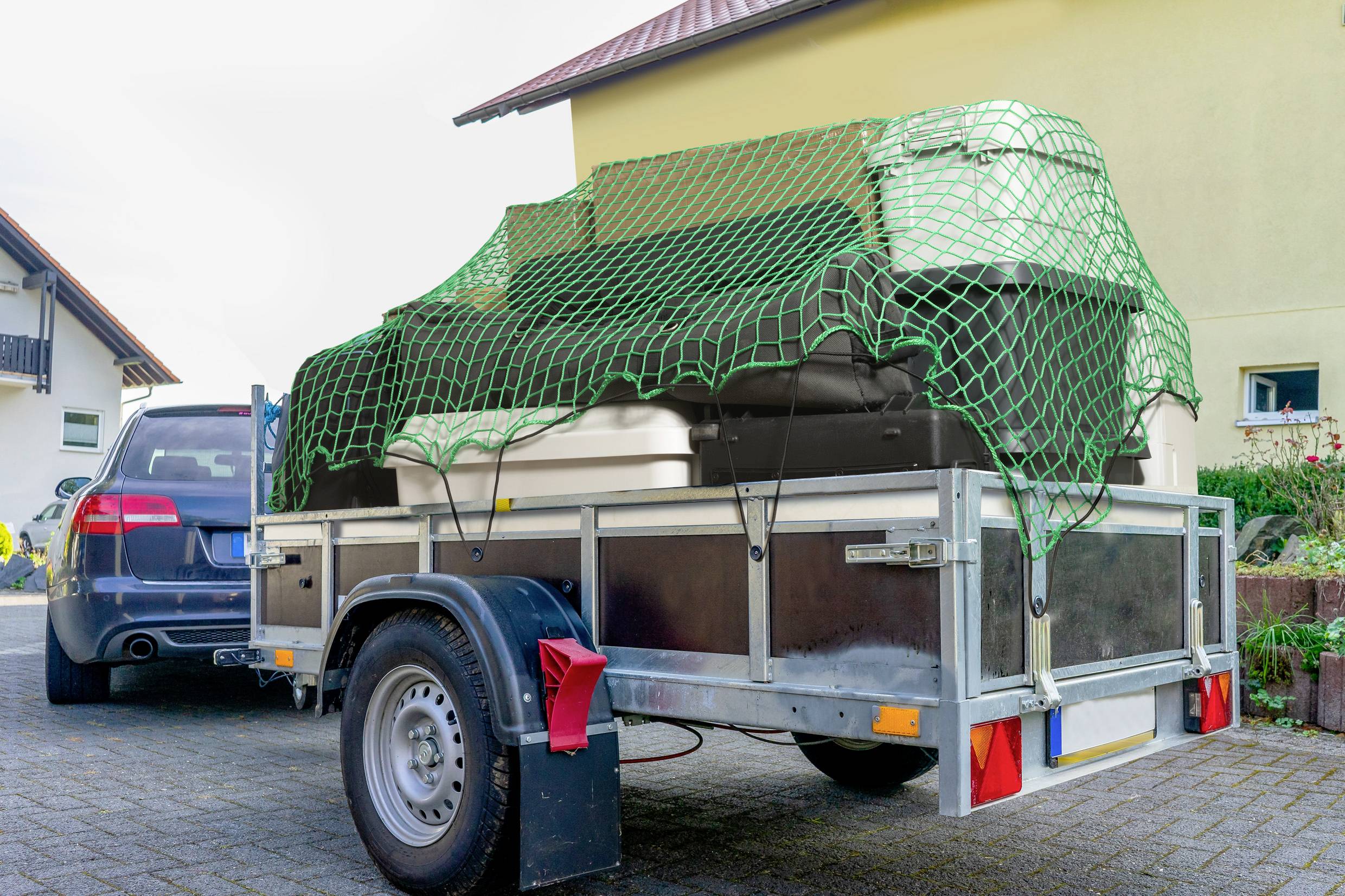 A car trailer, loaded with furniture and secured by a green net, is parked in a driveway next to a blue vehicle.