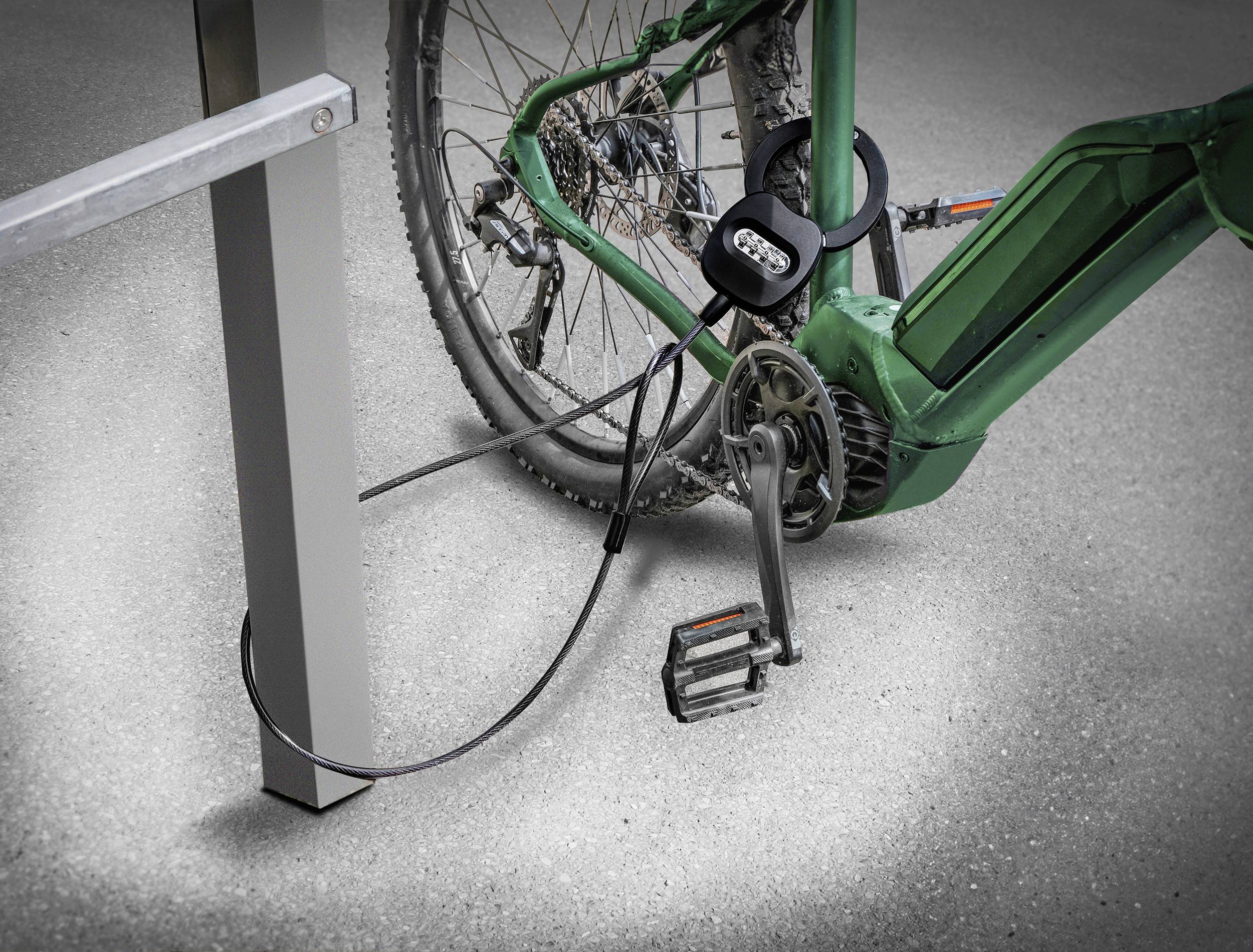 A green bicycle is secured with a black cable lock to a metal stand. The focus is on the lock and the rear wheel.