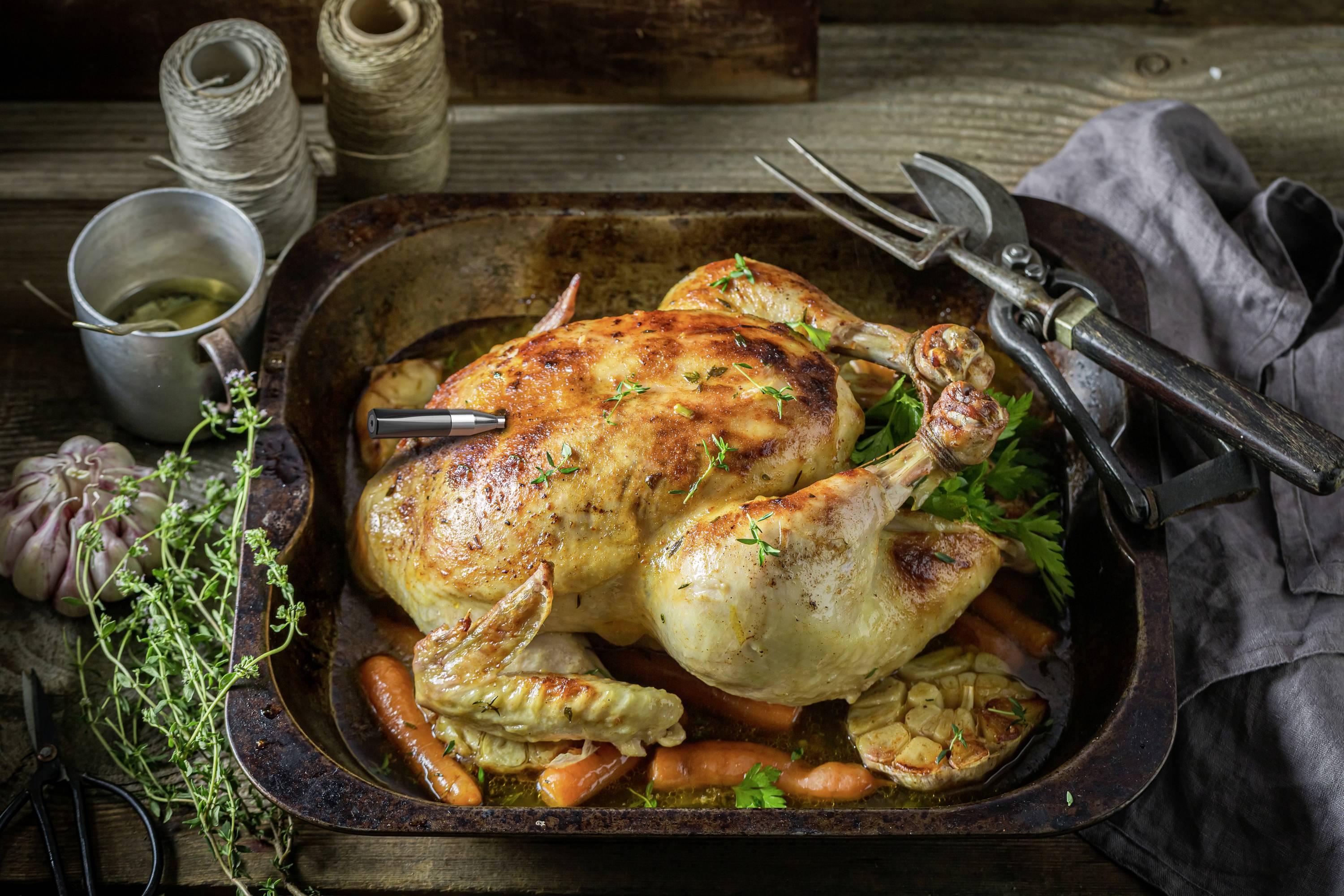 A roasted chicken breast in a pan surrounded by vegetables such as carrots and garlic, seasoned with fresh herbs.