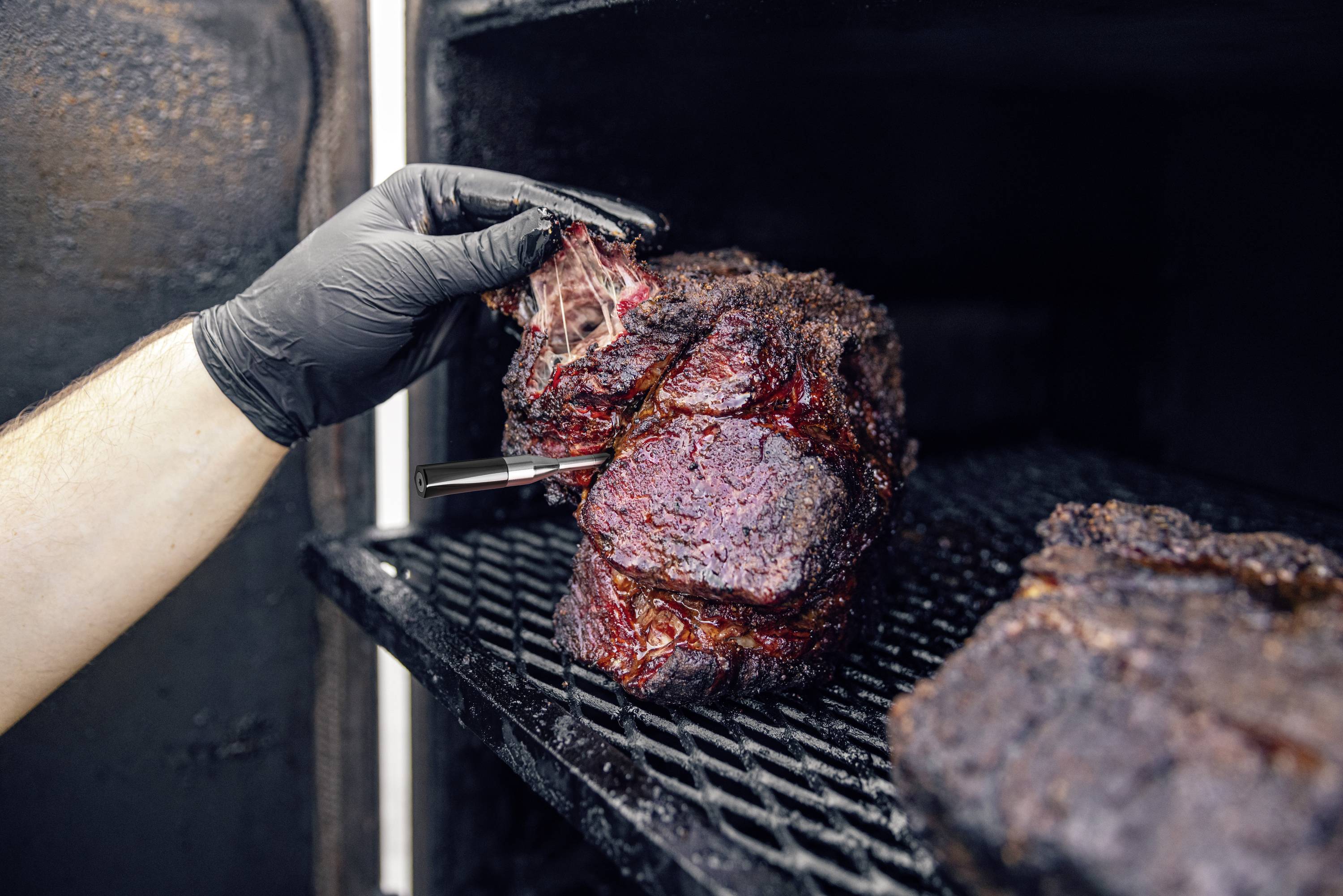 A hand in a black glove holds a large, smoked piece of meat with a thermometer. It is inside a smoker.