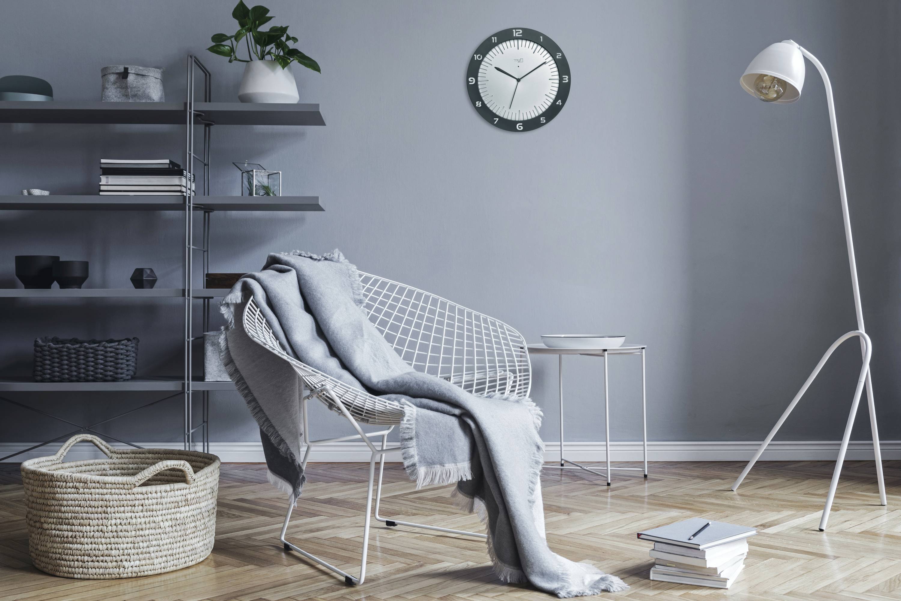Living room with a modern armchair, grey throw and lamp. A clock hangs on the wall. Shelving with plants and decorations in the background.