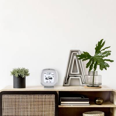 A shelf with a digital clock, plants, and decorative letters on a smooth surface. Against a white wall in the background.