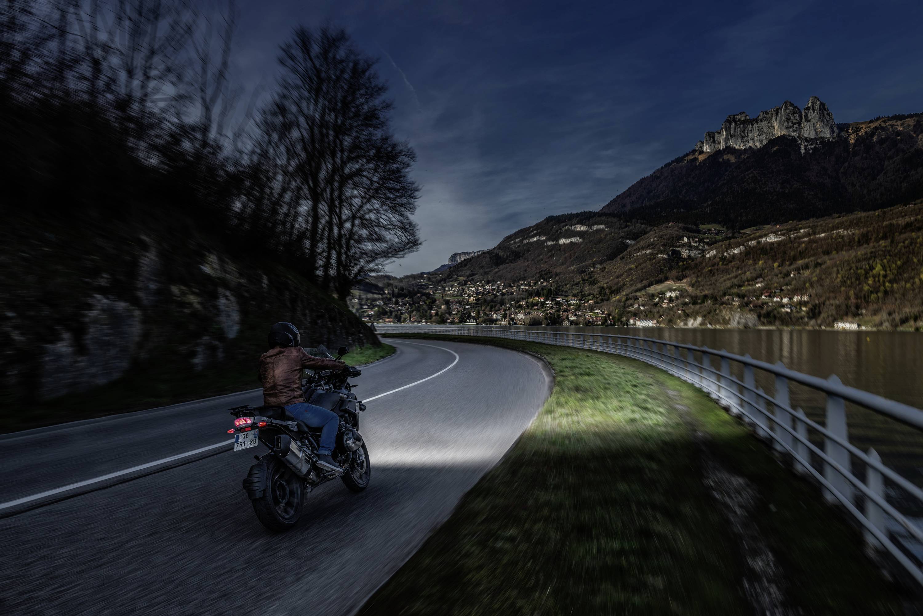 A person is riding a motorbike at night along a winding road beside a lake, surrounded by mountains in the background.