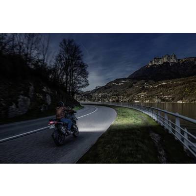 A person is riding a motorbike at night along a winding road beside a lake, surrounded by mountains in the background.
