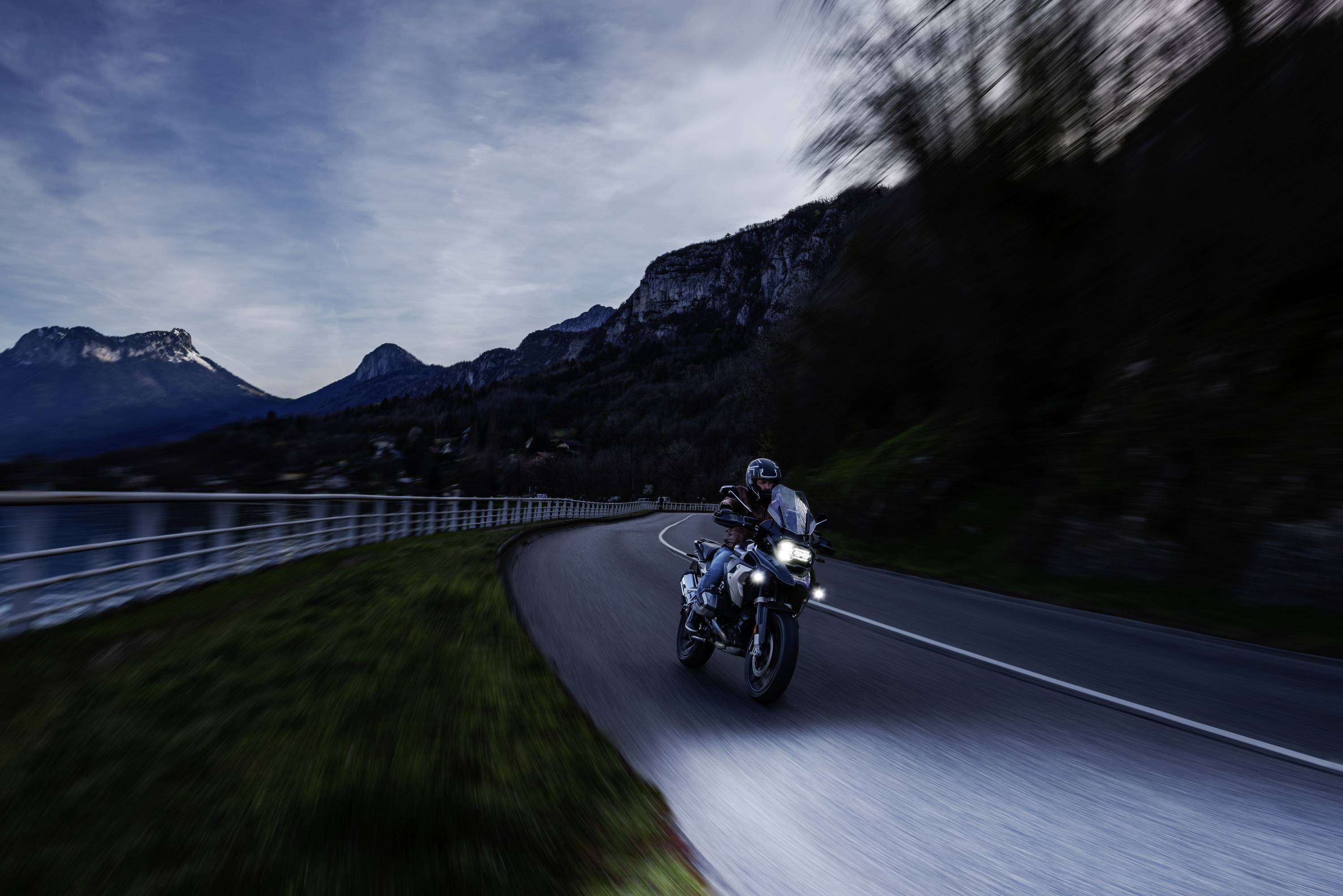 A motorcyclist rides along a winding road by a lake at dusk, surrounded by mountains and a sparsely illuminated landscape.