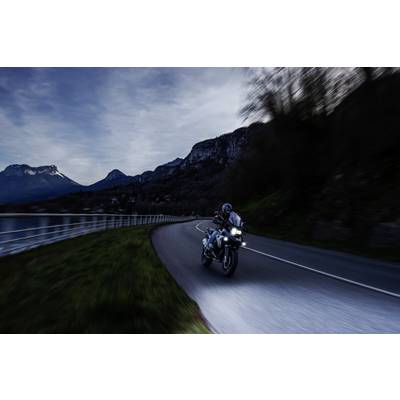 A motorcyclist rides along a winding road by a lake at dusk, surrounded by mountains and a sparsely illuminated landscape.