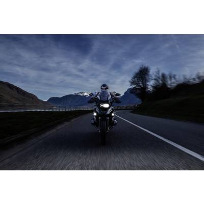 Motorcycle rider on a mountain road at dusk, headlights on, travelling towards the viewer. Mountains and trees in the background.
