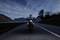 Motorcycle rider on a mountain road at dusk, headlights on, travelling towards the viewer. Mountains and trees in the background.
