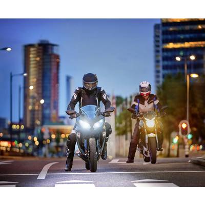 Two motorcyclists are riding on an urban street in the evening. They are wearing protective clothing and helmets. The background shows city buildings.