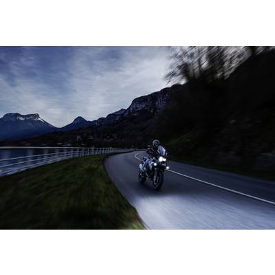 A person is riding a motorbike along a winding road at dusk, surrounded by mountains and a lake.