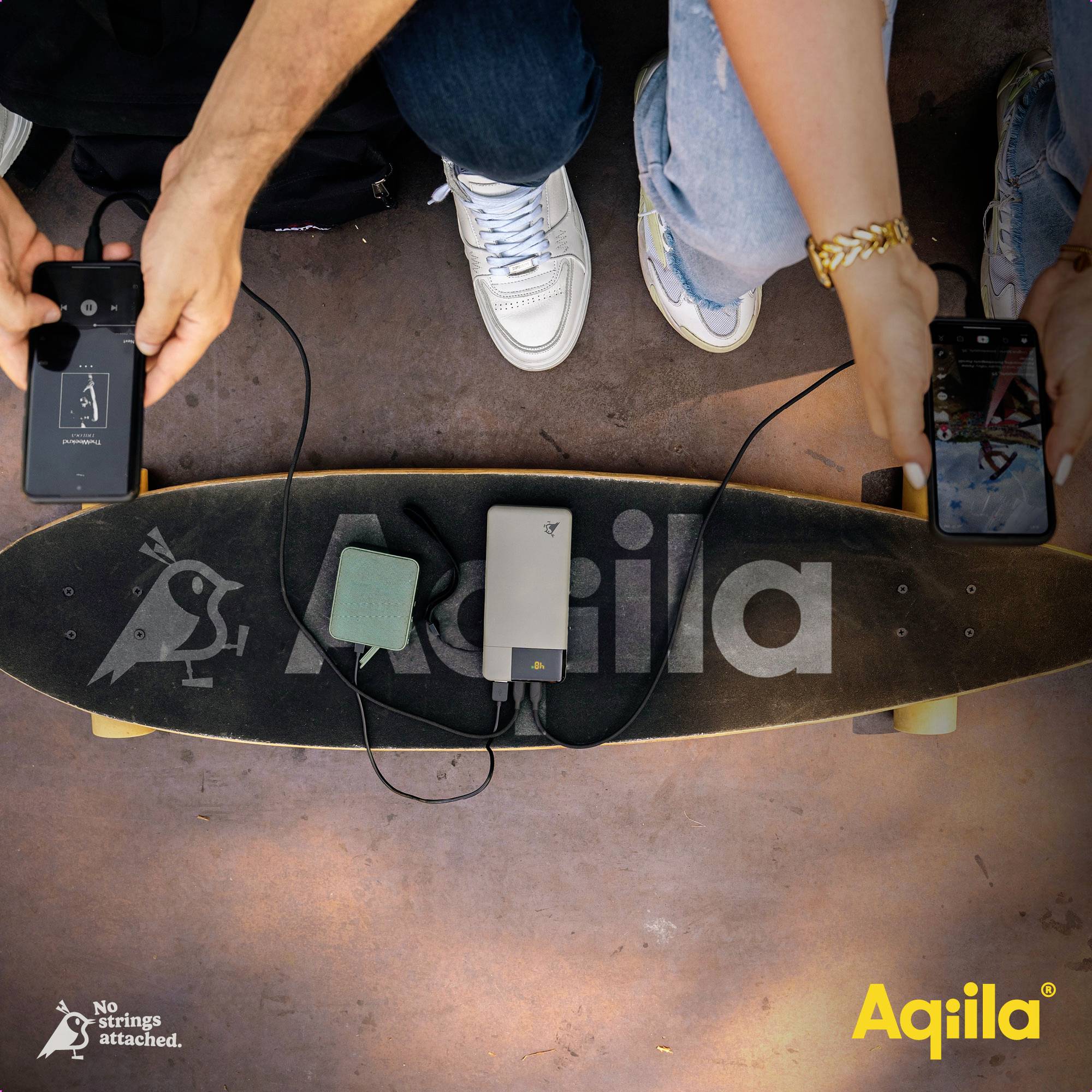Two people are sitting on a longboard. They are holding smartphones and looking at a charger lying between them. Text: 'No strings attached. Aqila'.<br><br>(Note: The translation is already in British English, so no changes were necessary.)