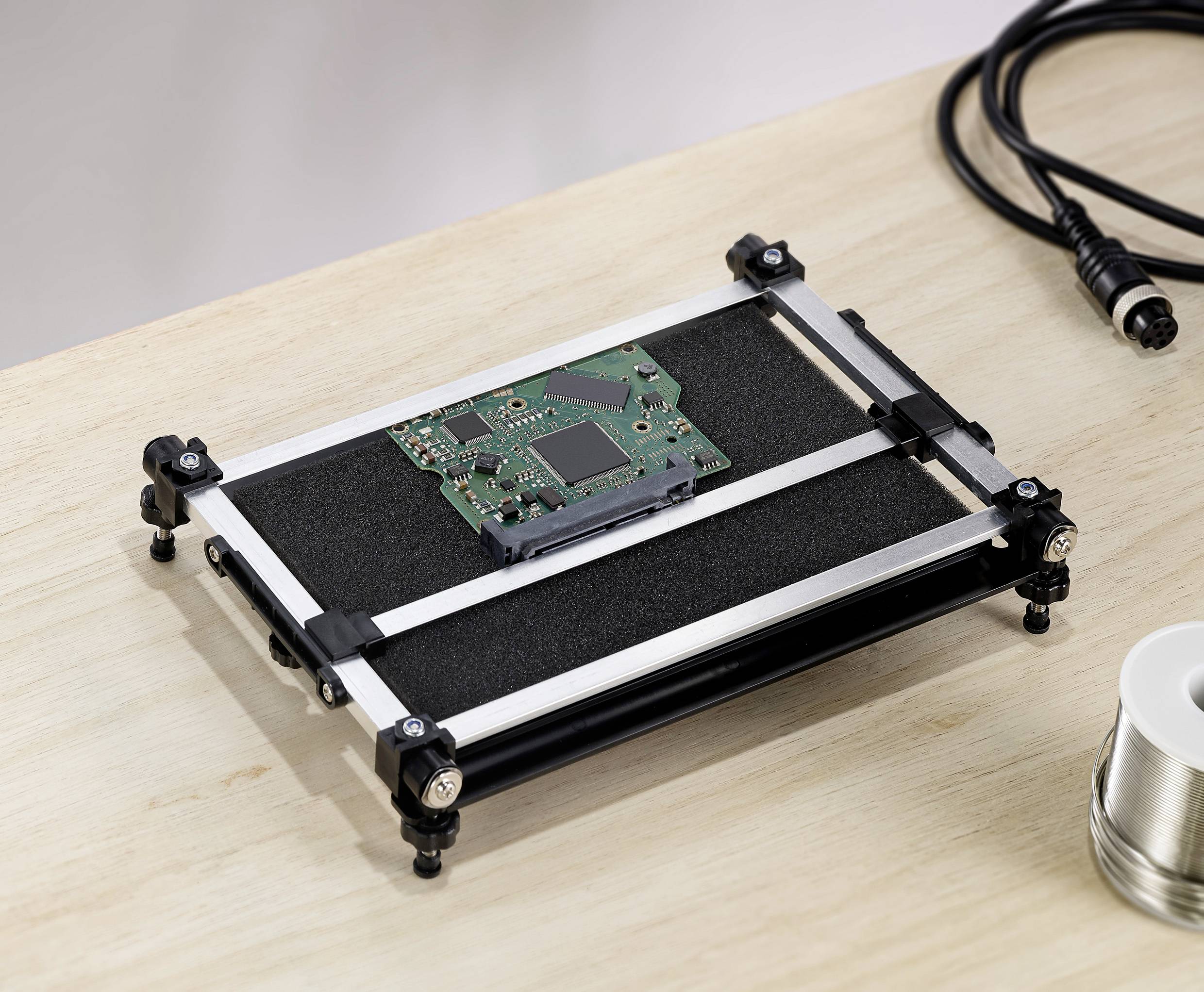 A green circuit board lies on an anti-static rack on a wooden table. Beside it, a cable and a reel of solder are visible.